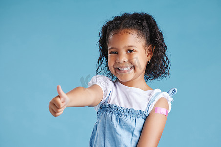 疫苗蓝色摄影照片_快乐的接种疫苗的孩子在注射疫苗后站在蓝色的工作室背景下，手臂上贴着石膏竖起大拇指。