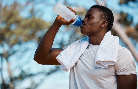 热毛巾摄影照片_年轻活跃的黑人男运动员在外面的森林里拿着一个瓶子，脖子上挂着毛巾喝水。