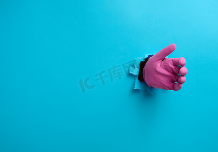 粉色乳胶手套里的一只手握着一个物体，身体的一部分从蓝纸背景中的一个破洞里伸出来