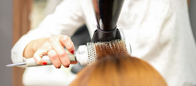 一位专业美发师正在用吹风机和圆刷烘干长红头发，特写。