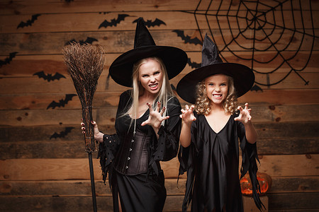 女孩与母亲合影摄影照片_万圣节概念-欢快的母亲和她的女儿穿着女巫服装庆祝万圣节，在木制工作室背景下与蝙蝠和蜘蛛网上的弯曲南瓜合影。