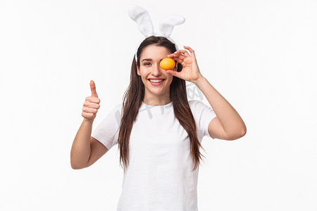 兔耳中满意、快乐的年轻女子的肖像，眼睛上拿着一颗彩蛋，竖起大拇指为复活节假日派对画画，庆祝传统活动，白色背景