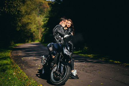 一辆摩托摄影照片_英俊的年轻人和美丽的年轻女孩，夫妇坐在摩托车上，站在一辆穿着黑色皮衣的摩托车附近，拥抱，在大自然中，在街上