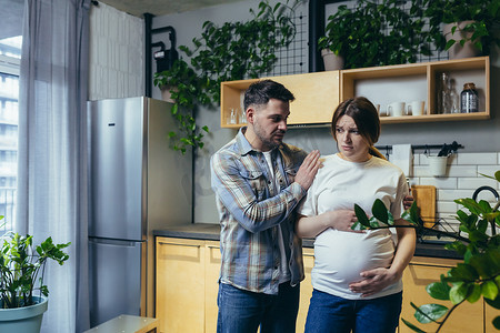 怀孕悲伤摄影照片_年轻家庭的男人和怀孕的妻子、丈夫在厨房争吵后安抚女人