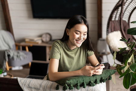 女孩购物摄影照片_微笑的韩国女孩用手机聊天，坐在家里的客厅里，拿着智能手机，笑得很开心