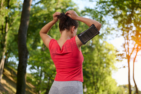准备跑步的人摄影照片_一位戴着智能手机支架、扎马尾辫、准备沿着城市公园户外跑步的女运动员的后视图