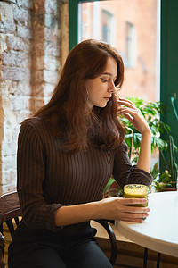 漂亮、严肃、时尚、时髦的聪明女孩坐在咖啡馆的窗边，喝着健康的黄色冰沙或素食拿铁。