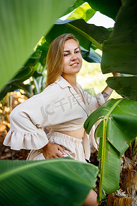 在热带地区的一个公园里，靠近香蕉灌木绿叶的女人，她穿着米色裙子和衬衫