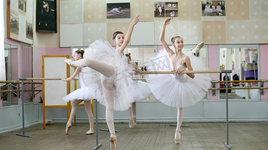 在芭蕾舞厅，穿着白色芭蕾舞裙的女孩正在芭蕾舞中排练姿势，年轻的芭蕾舞演员穿着足尖鞋站在脚趾上，在芭蕾舞厅的栏杆上优雅地向后抬起双腿。
