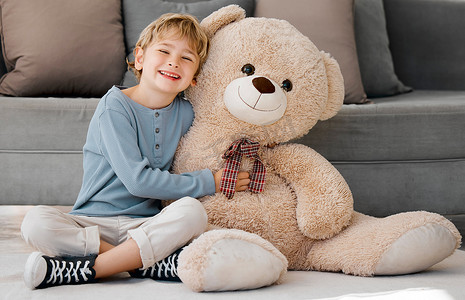 他在家摄影照片_一个快乐的白人小男孩的肖像，他在家里的休息室的地板上抱着一只又大又舒适的毛茸茸的泰迪熊微笑着。