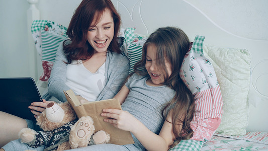 童话书摄影照片_可爱的小女儿和年轻慈爱的母亲一起坐在家里舒适的床上，看着有趣的图片，笑着读童话书