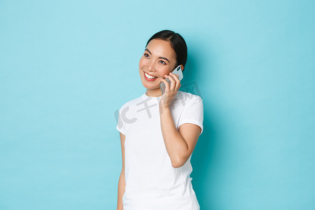微笑的现代亚洲女孩在打电话时看起来很高兴的肖像，确认在线订单，与朋友聊天，站在浅蓝色背景。