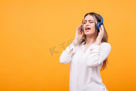 黄色背景下用蓝色耳机唱歌和听音乐的兴奋女孩的肖像