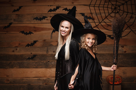 女孩与母亲合影摄影照片_万圣节概念-欢快的母亲和她的女儿穿着女巫服装庆祝万圣节，在木制工作室背景下与蝙蝠和蜘蛛网上的弯曲南瓜合影。