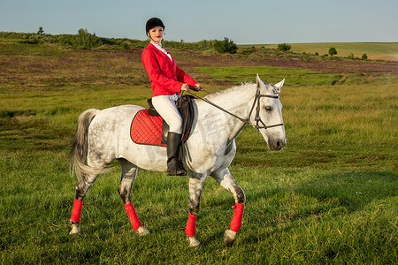 女骑手摄影照片_年轻的女骑手，穿着红色的 redingote 和白色的马裤，她的马在夕阳的余晖下。