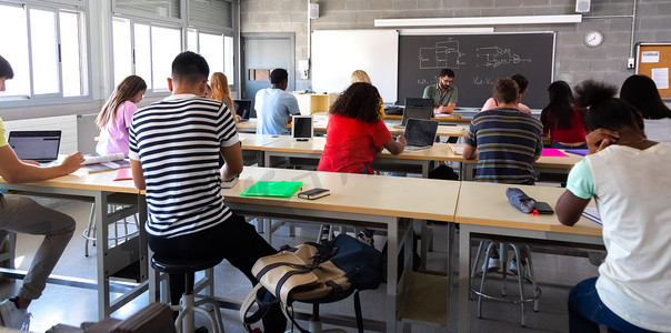 当老师批改考试时，一群多种族高中生在课堂上使用笔记本电脑的后视图。