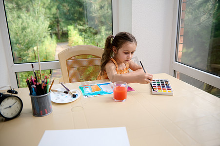 白人儿童，小学年龄的女孩坐在桌子旁，用水彩和画笔画画。