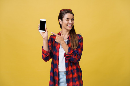 一个穿着夏季服装的快乐年轻女孩的肖像，手指着黄色背景中突显的空白屏幕手机
