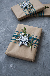 三件用牛皮纸包裹的圣诞礼物，蓝色线包裹着，灰色混凝土背景上有白色雪花和迷迭香枝条。