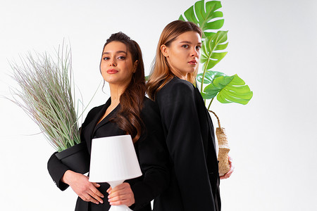 两个穿着黑色西装的年轻女商人微笑着拿着室内植物和孤立在白色背景上的灯。