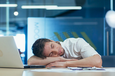 年轻疲惫的商人睡在办公室工作场所的桌子上