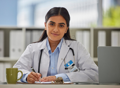 一位戴着听诊器的年轻印度医生的画像坐在办公室里，她坐在办公桌前开处方。