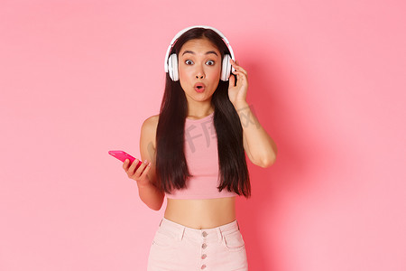 惊讶而兴奋的亚洲可爱女孩盯着相机，一边听有趣的播客或朋友播放列表，一边用耳机听新歌，一边拿着手机、粉色墙壁，一边好奇