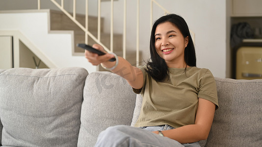 快乐的年轻女人拿着遥控器，在沙发上看电视，在家享受无忧无虑的休闲周末活动