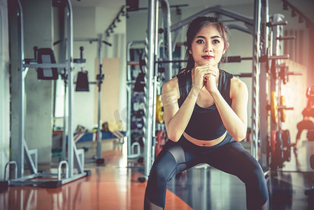燃烧脂肪摄影照片_年轻的亚洲女性在背景有运动器材的健身运动馆里进行蹲式锻炼，以燃烧脂肪和节食。