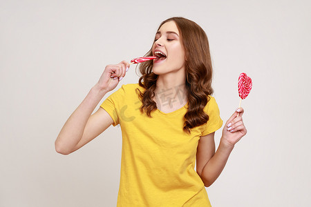 穿着黄色休闲 T 恤的女人肖像咬棒棒糖，带着满意的表情品尝甜心形糖果，享受美味的风味甜点。