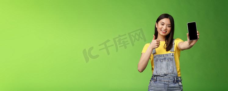 开朗可爱的亚洲女性判断优秀的智能手机游戏，展示大拇指标志，伸出手臂握住电话屏幕，展示出色的应用程序，推广应用程序，站立绿色背景