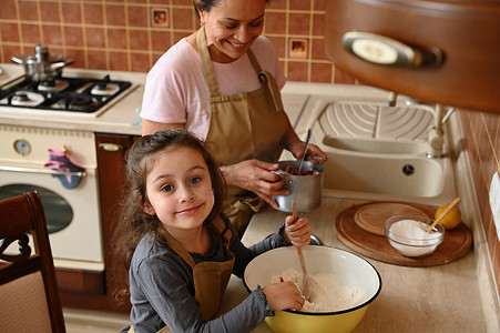漂亮的孩子，穿着厨师围裙的欧洲小女孩，站在妈妈旁边的厨房台面旁揉面团