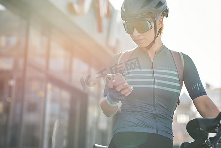 女自行车运动员摄影照片_在市中心骑自行车时使用智能手机的防护装备的女自行车手。