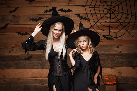 万圣节概念-欢快的母亲和她的女儿穿着女巫服装庆祝万圣节，在木制工作室背景下与蝙蝠和蜘蛛网上的弯曲南瓜合影。