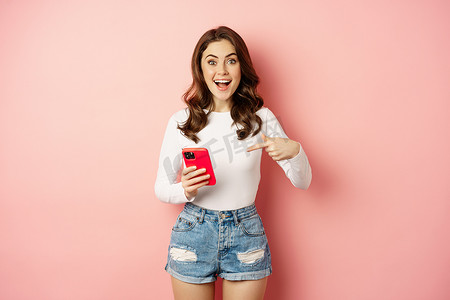 快乐的黑发女孩发布公告，指着手机，展示在线促销、应用新闻，站在粉红色背景下
