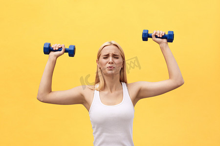 照片中，严肃的年轻运动员用黄墙背景上的哑铃进行锻炼。