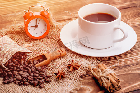 木质背景中的一杯咖啡，配有肉桂条、茴香、咖啡豆和红色闹钟