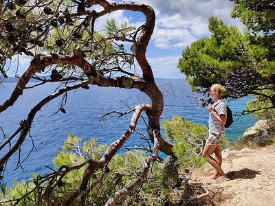 年轻活跃的 feamle 游客背着小背包走在松树间的沿海小路上，寻找偏远的海湾，在克罗地亚的海边安静地游泳。
