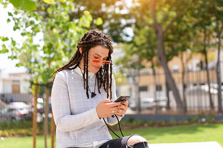 戴着粉色太阳镜、留着长发绺的年轻嬉皮士使用智能手机，坐在户外的夏季公园里。