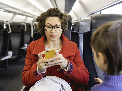 短卷发男孩摄影照片_穿着红色粗呢大衣的卷发女人在郊区火车上用智能手机发短信。