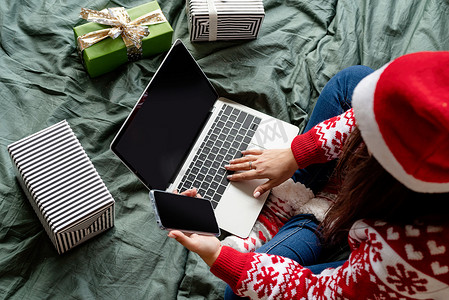 穿着圣诞衣服的女人坐在床上用笔记本电脑和手机