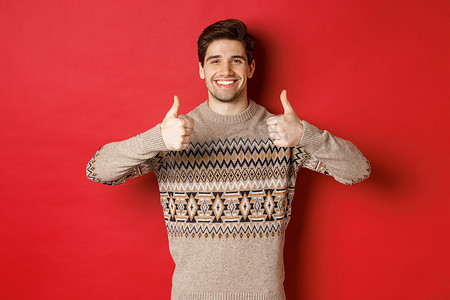 穿着圣诞毛衣的快乐而高兴的帅哥的肖像，竖起大拇指，点头表示赞同，满意地微笑，站在红色背景上