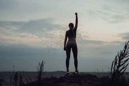 运动女孩站在山顶并在晚上举起手的剪影，女性力量