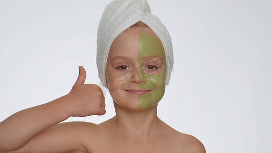 头上戴着毛巾的可爱微笑小女孩，脸上涂着清洁保湿绿色面膜