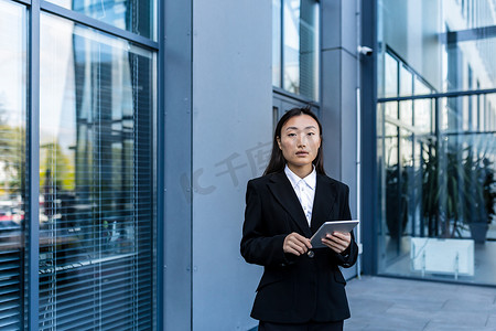 身穿商务正装的亚洲女商人站着，手里拿着平板电脑工作