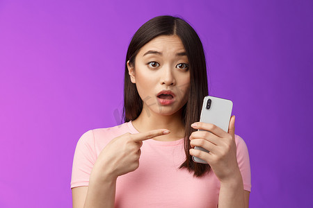 忧心忡忡的困惑可爱的亚洲年轻女性拿着智能手机张开嘴，指着电话，看起来很沮丧，在网上看到奇怪的帖子，站在紫色背景上