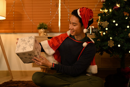 家庭欢庆摄影照片_戴着圣诞帽的微笑男子打开圣诞礼物，坐在装饰精美的房间里庆祝新年和圣诞佳节