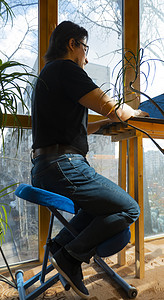 人体工程学摄影照片_一名男子坐在阳台上的符合人体工程学的矫形跪椅上使用笔记本电脑工作。