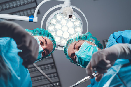 医疗帽子摄影照片_在手术室明亮的灯光下，身着医疗制服的女外科医生使用专业工具站在下方