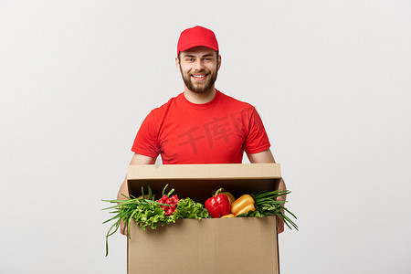蔬菜超市摄影照片_送货概念：英俊的白人杂货送货快递员穿着红色制服，配有新鲜水果和蔬菜的杂货箱